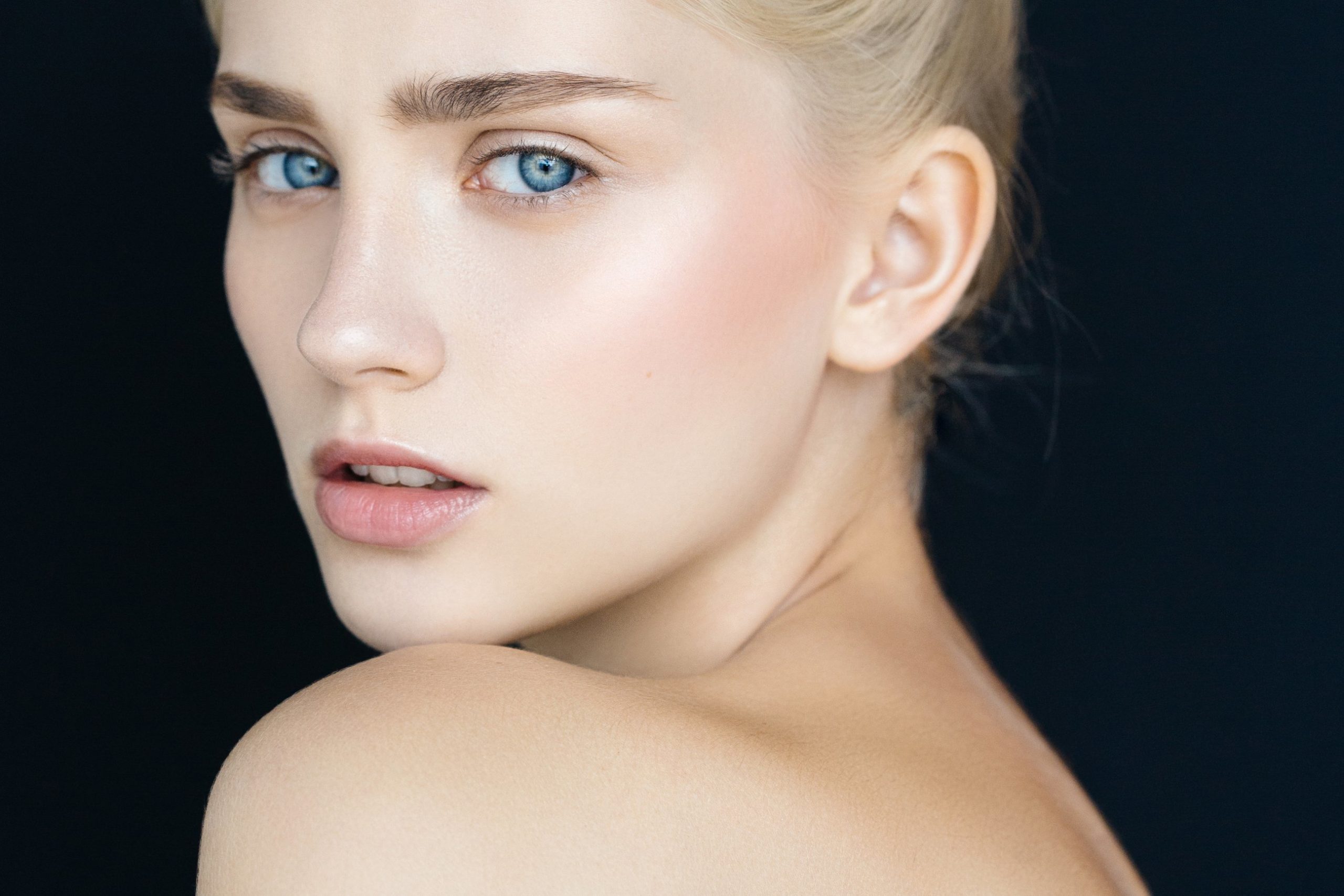 Close-up portrait of a young woman with glowing skin, blue eyes, and bare shoulders against a dark background.